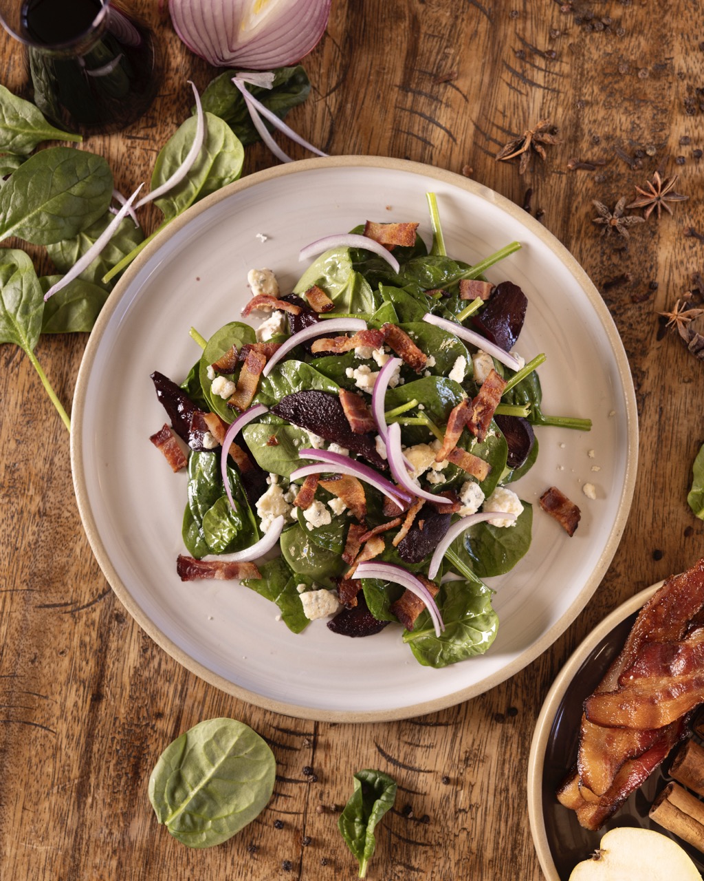 A salad sits on a white plate on top of a wood table has spinach leaves surround the plate.