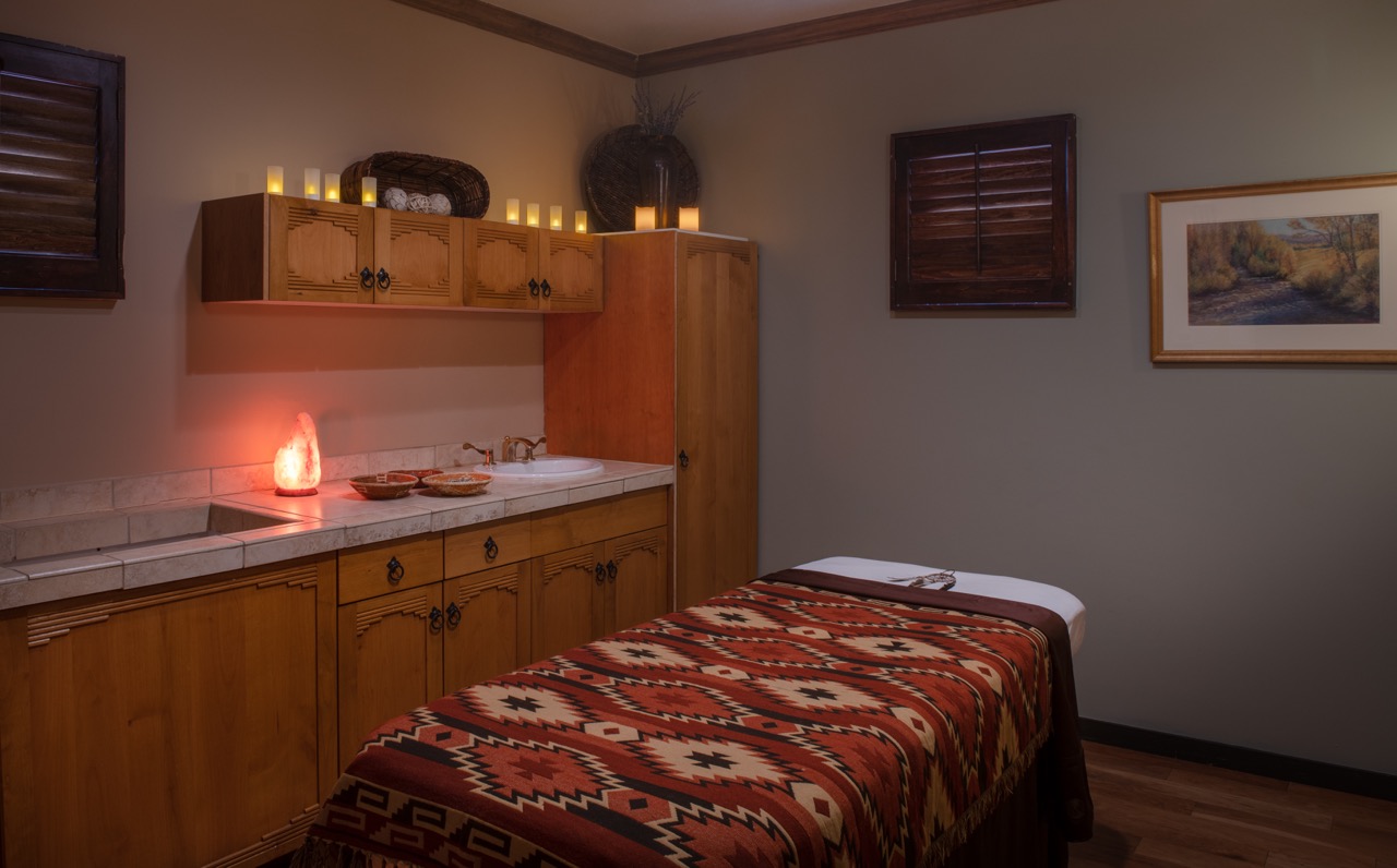 A small red massage bed sits in the center of a spa room with candles on a counter nearby.