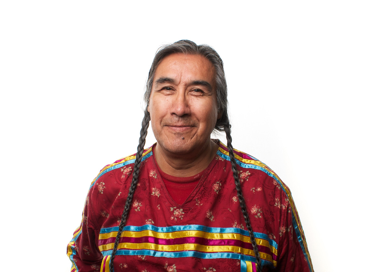 A Native American man with two long braids in a red shirt on a white background