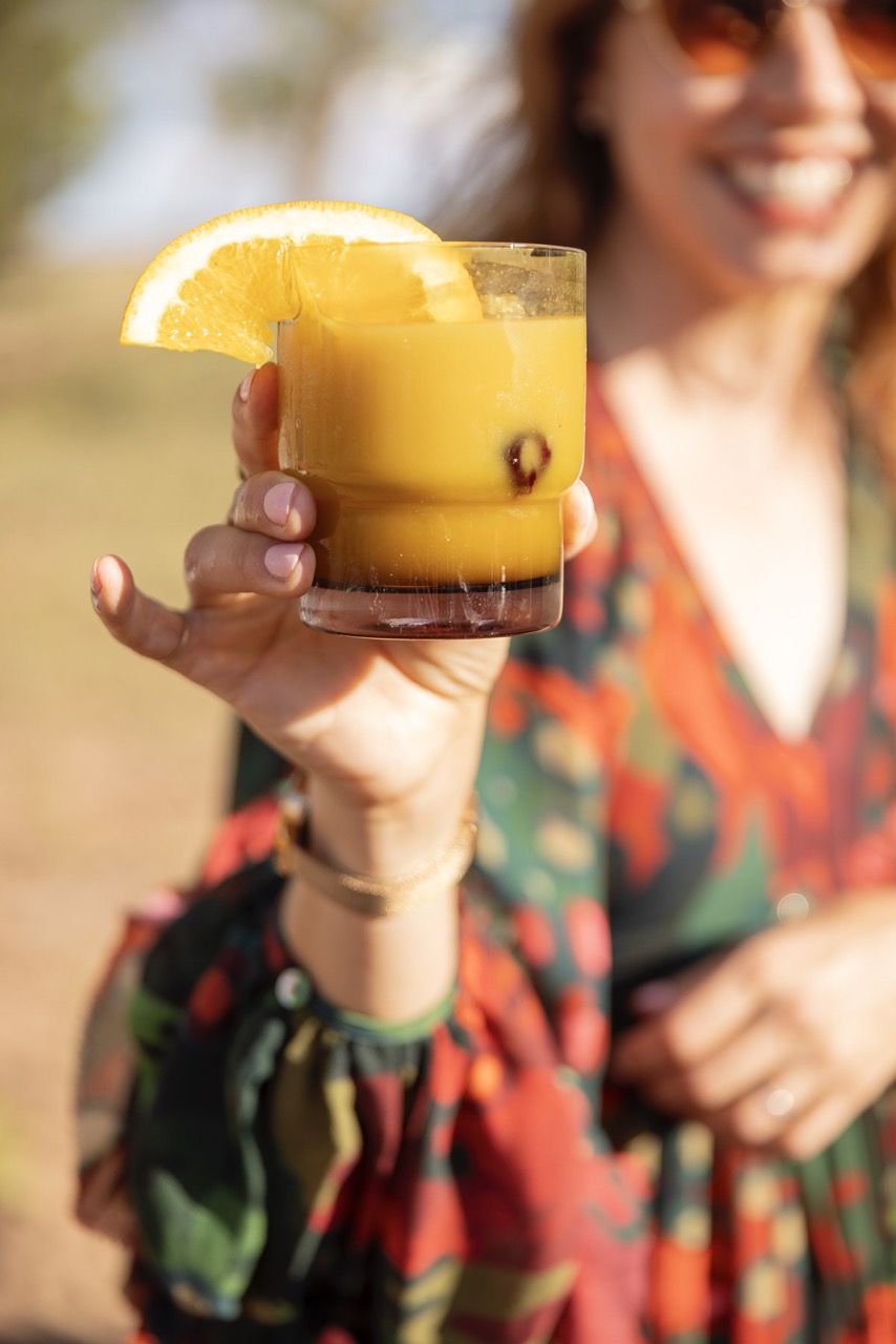 A woman holds up an orange cocktail with an orange slice in it. 