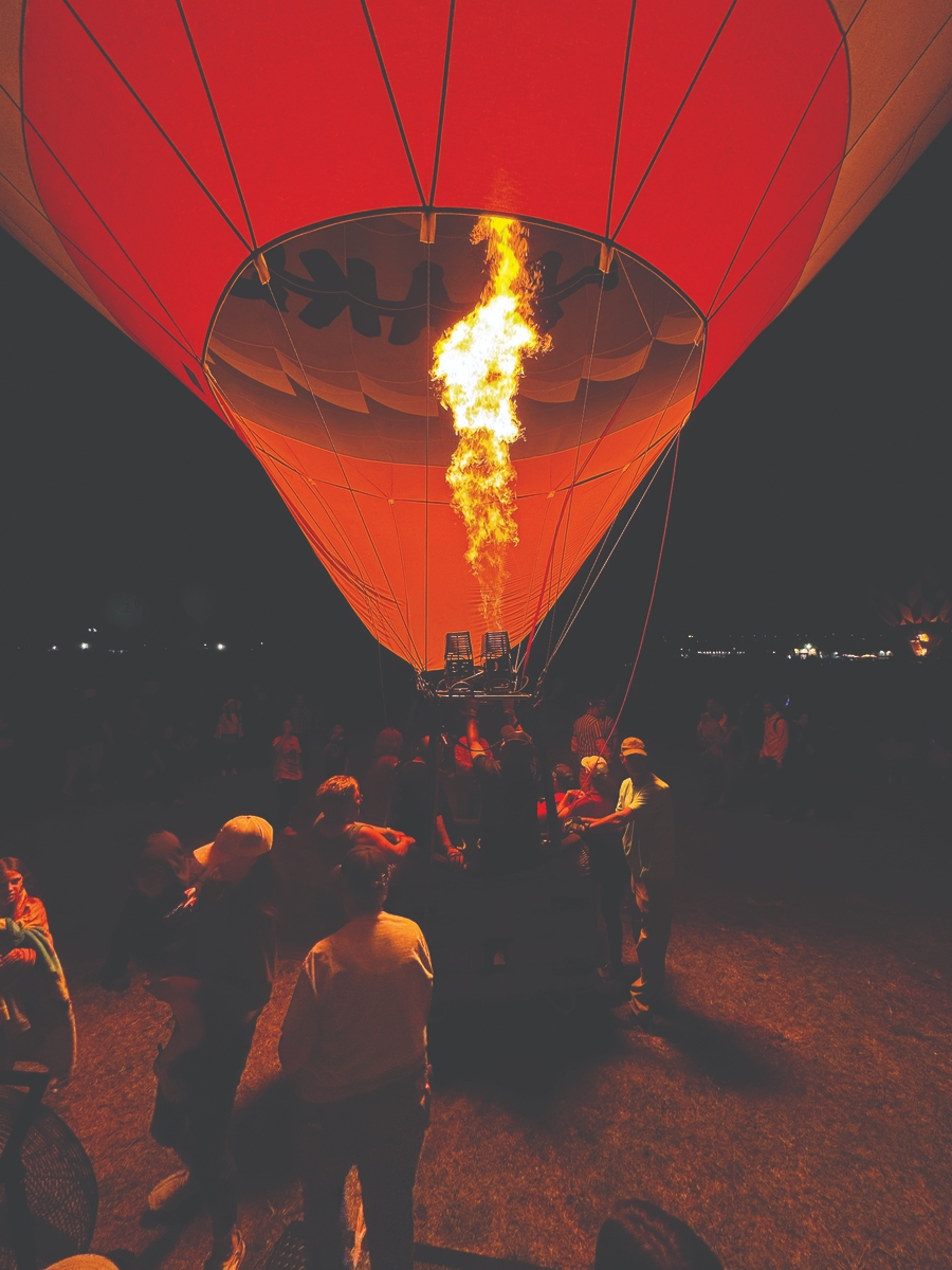 Hot air balloon glowing with flame at night during a festive event, surrounded by an enthusiastic crowd.