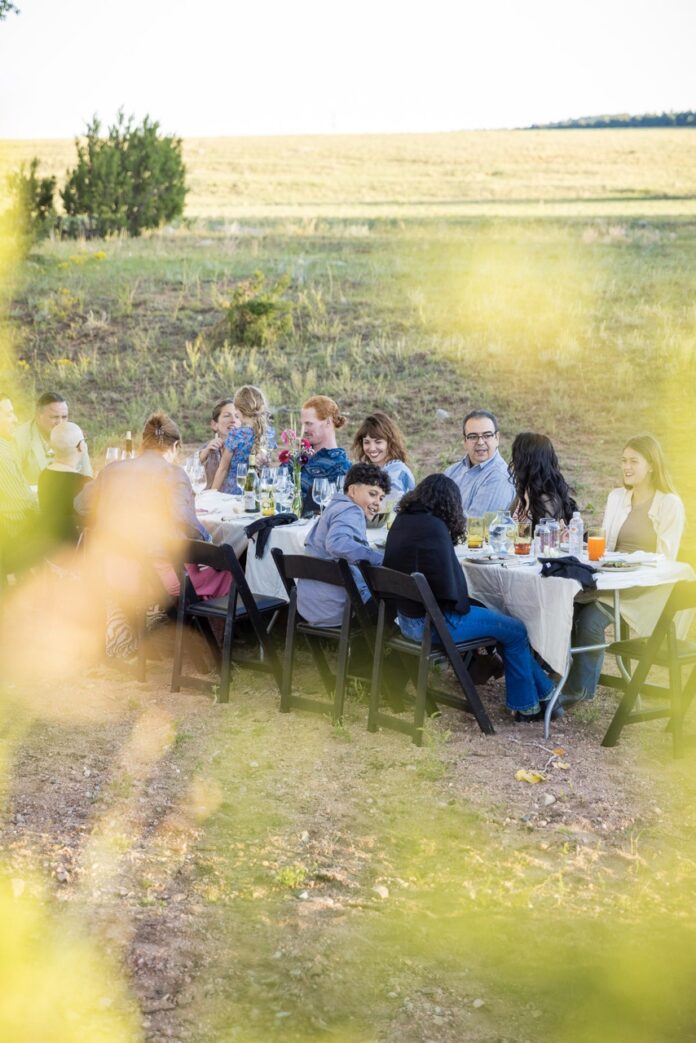 A group of people sit at a Beck and Bulow dinner at a white table as tree branches obstruct a clear view.