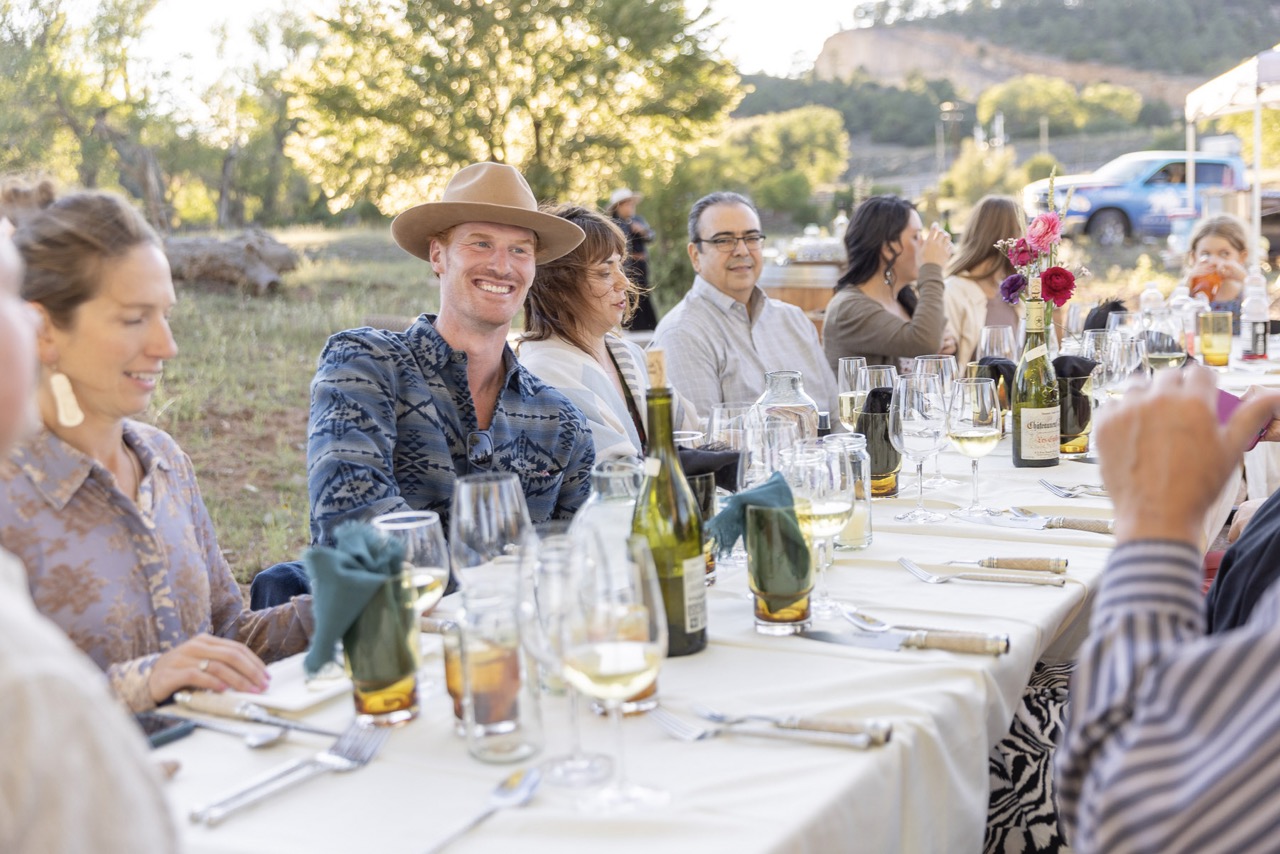 A group of people at a white table smile for a photo with wine bottles amongst the table.