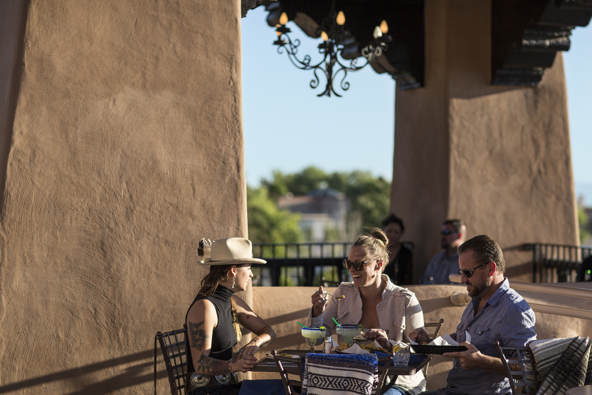 Outdoor dining scene at La Fonda hotel's rooftop Bell Tower bar in Santa Fe, showing three guests casually enjoying a meal under the sun with iconic views of the city's historic landmarks in the background.