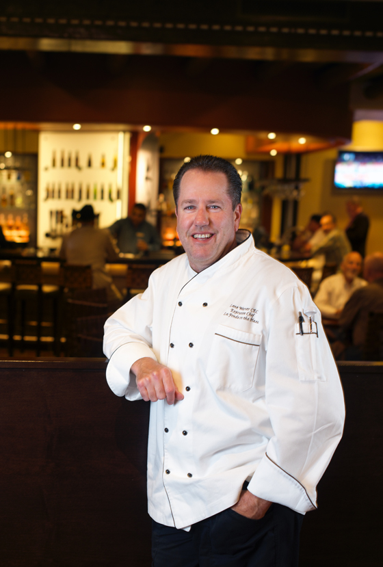 Chef Lane Warner standing in a modern commercial kitchen, wearing a white uniform and gesturing while speaking to others present.