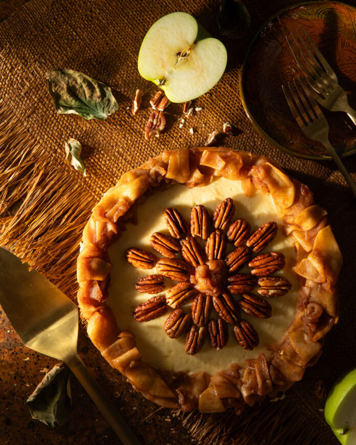 Sliced apples arranged artistically around a cream cheese based cheesecake drizzled with cinnamon sitting atop a crushed shortbread crust and topped with pecan pieces.