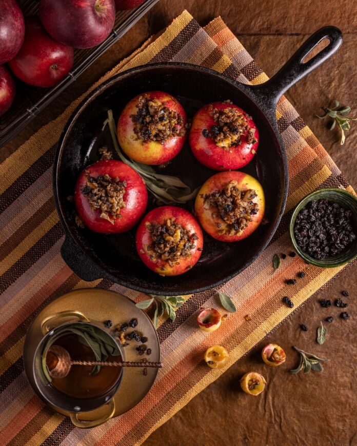 Baked apples topped with melted cheese and spices sitting on a wooden surface with scattered apple slices and rosemary nearby.
