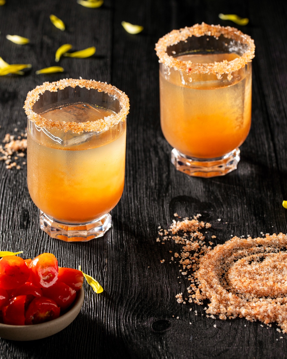 Two golden cocktails topped with a coarse salt rim and berry-colored sprinkles in short rocks glasses on a dark wooden surface, featuring the 'Lyrical Thesis' cocktail crafted with agave spirits at Happy Accidents.