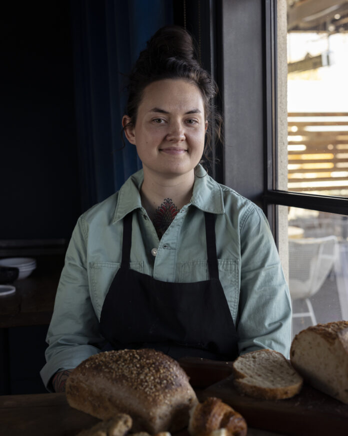 A woman in a green apron standing in front of a table with various baked goods on display, including bread and pastries.