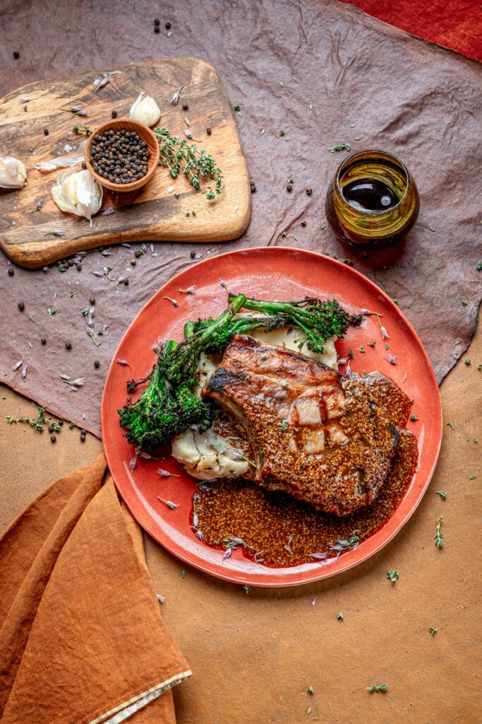 Grilled pork chop with whole grain mustard demi, broccolini, and roasted garlic mashed potatoes on a rustic plate.