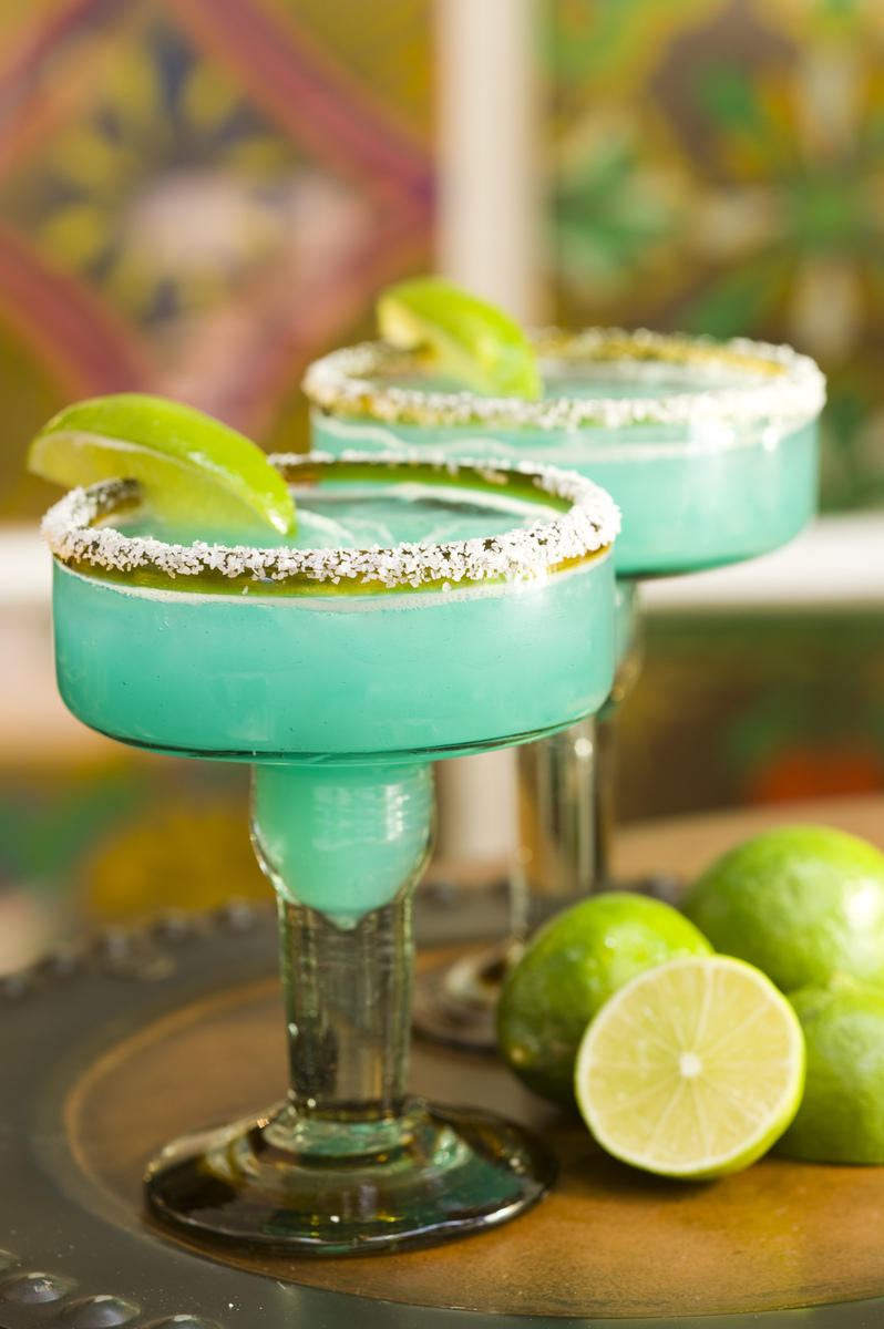 Two lime green margarita cocktails in stylish glassware laying on top of a wooden bar, expertly garnished with lime wedges and salt-dusted rims for an inviting presentation.