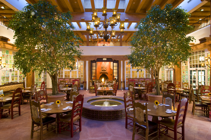 Interior dining area at La Plazuela restaurant inside La Fonda hotel in Santa Fe, showing tables dressed with white linens and floral centerpieces beneath hanging string lights, creating an ambient Southwestern atmosphere.