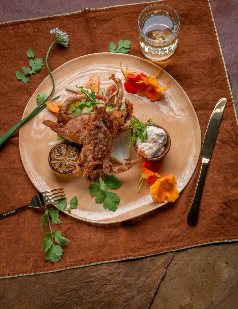 Crispy tempura soft shell crab on a ceramic plate with lemon, herbs, and dipping sauce, elegantly presented.