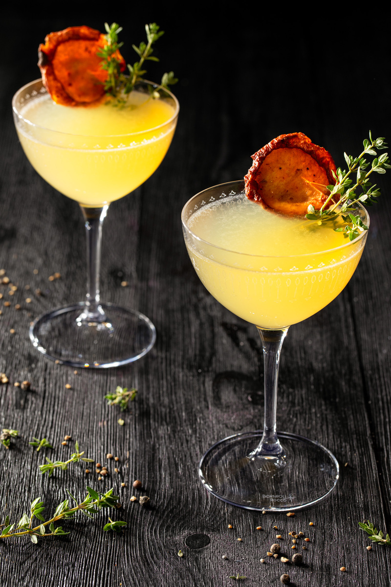 Two yellow cocktails garnished with apple chips and thyme sprigs in tall coupe glasses sitting on a wooden surface surrounded by scattered herbs and spices.