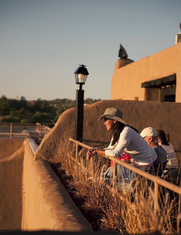 A lone individual seated on an outdoor bench at night, gazing downward in quiet contemplation with the luminous skyline of Santa Fe in the distance.