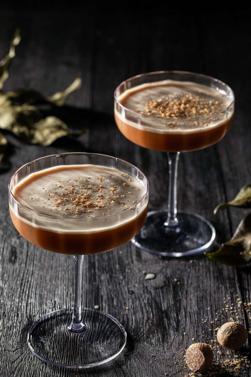 Two whiskey colored cocktails garnished with grated nutmeg in tall coupe glasses sitting on a wooden surface featuring the fall-inspired 'Solstice Sour' cocktail.