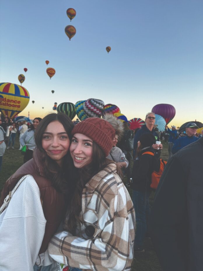 Two friends smiling at the Albuquerque International Balloon Fiesta with colorful hot air balloons in the background.