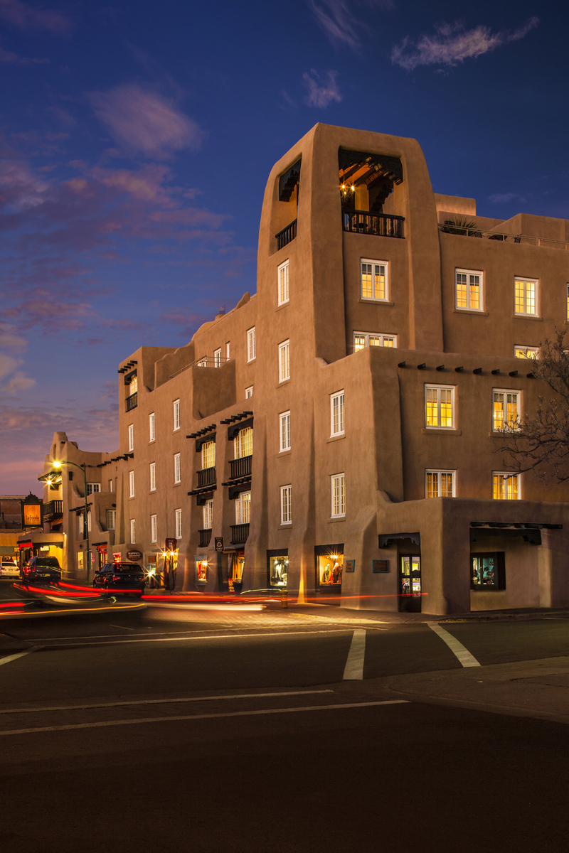 Panoramic view of La Fonda hotel silhouetted against a vibrant orange and pink skyline at dusk, as the lights of the city of Santa Fe gradually illuminate below.