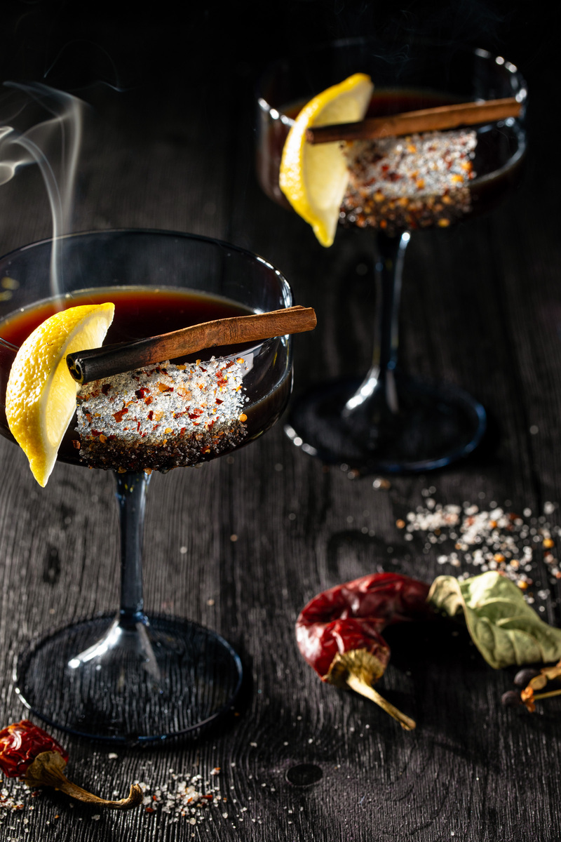 Two rustic amber-colored drinks garnished with cinnamon sticks and lemon wedges in rocks glasses sitting on a wooden surface scattered with baking spices, featuring the fall-inspired 'Tamarind Sour' cocktail.