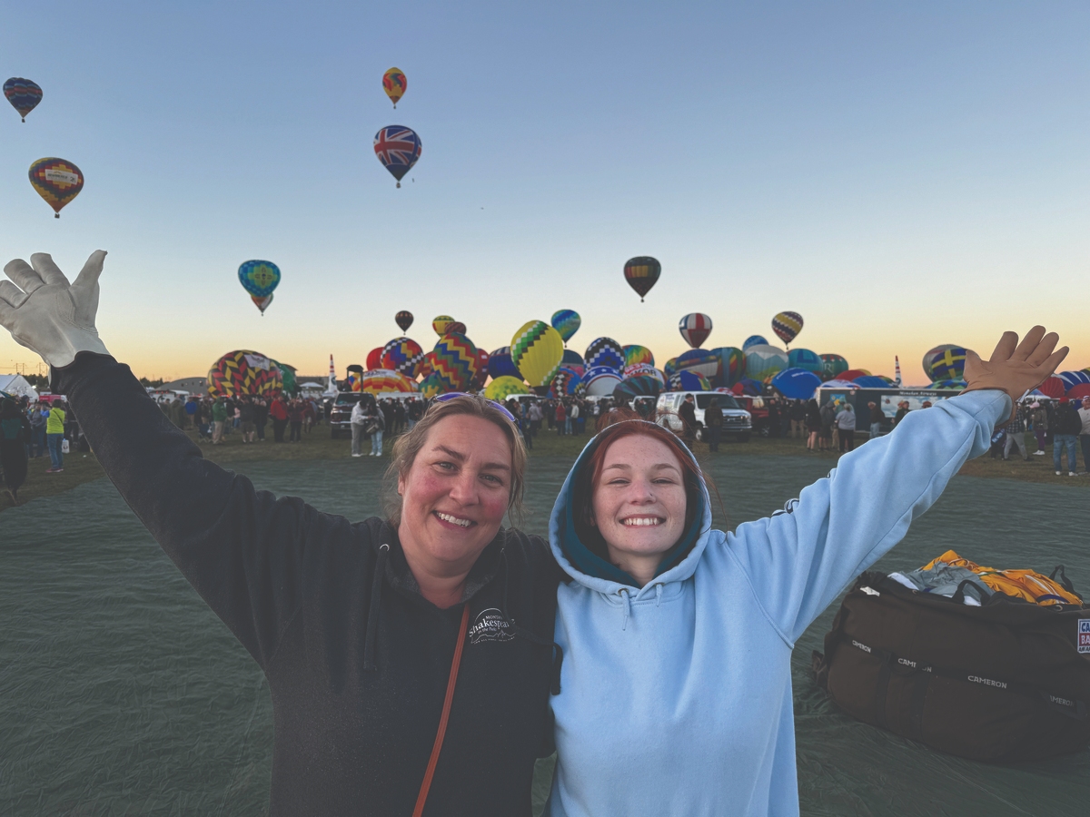 Two women joyfully celebrating at the Albuquerque International Balloon Fiesta with colorful hot air balloons in the background.