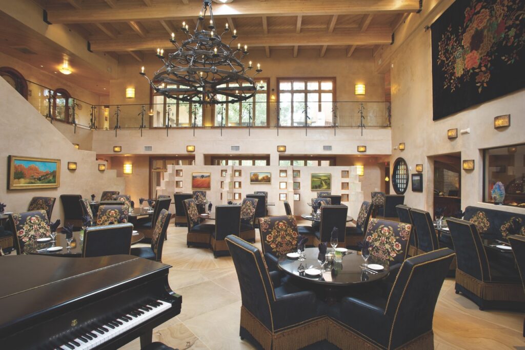 A Winter Staycation at El Monte Sagrado An indoor dining room with black tables and chairs, a black chandelier and a piano.