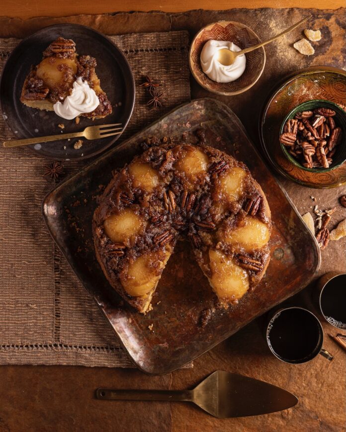 A pear upside down cake sits on a wooden table with a slice taken out of it on a plate to the top left.