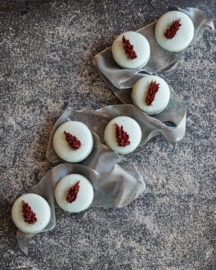 A line of blue corn macarons with red floral pieces on tops sit on a silver ribbon on a grey table.