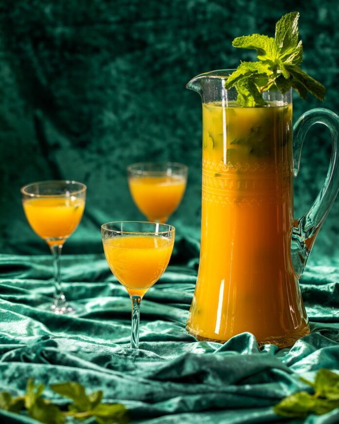 Piña Paradise Mojito, A Batched Cocktail A large glass pitcher filled with a vibrant orange Piña Paradise Mojito cocktail garnished with mint, beside two glasses ready to serve.