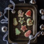 A few holiday themed Iced Sugar Cookies sit on a metal tin tray with ribbon and ornament bulbs around the try.