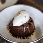 A piece of Malva Pudding sits in a white bowl dish with a scoop of white cream on top and nuts.