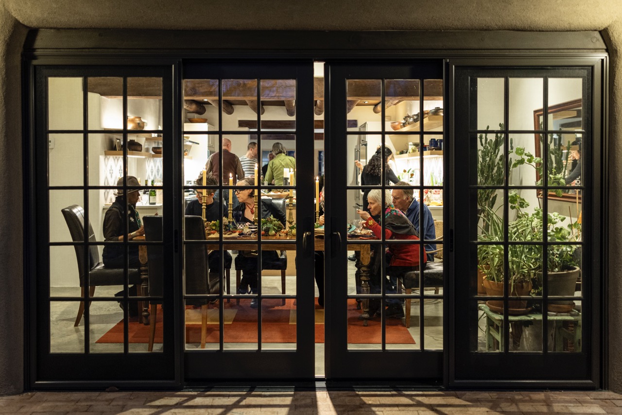 A group of people sit and eat at a dining room table as they're framed through window doors. 