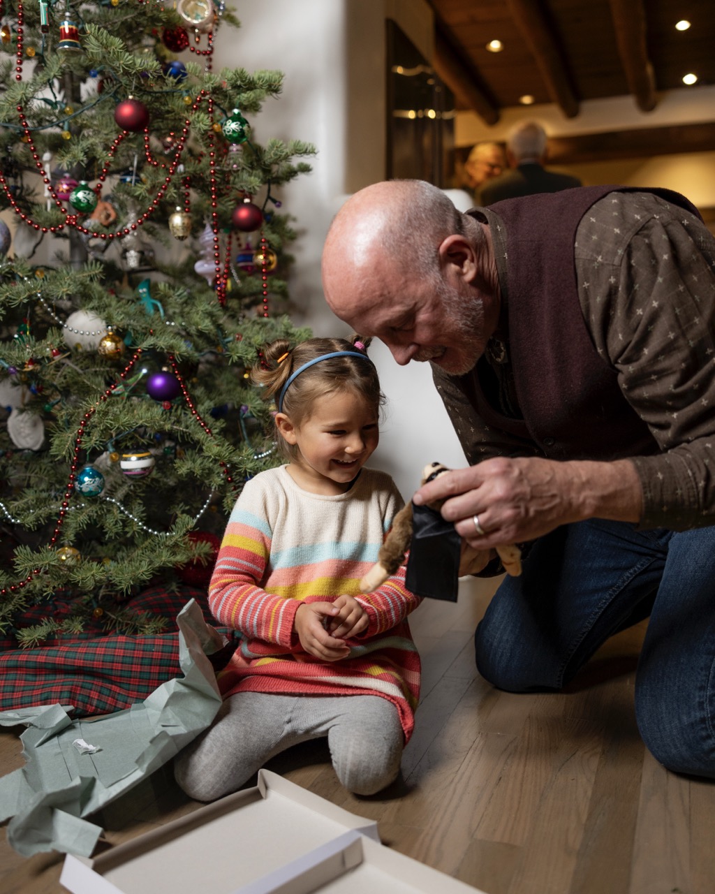 An older man and a little girl play with a toy near the Christmas tree.