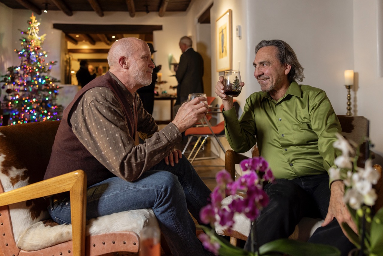 Two men smile at each other in conversation at a Christmas party with drinks in hand.