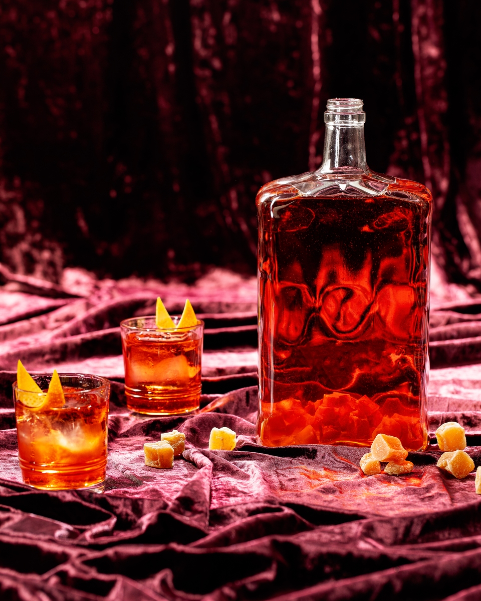 A glass filled with an amber colored cocktail garnished with a candied ginger slice, illustrating the "Starlight Negroni" batch cocktail.