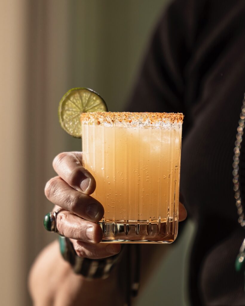 A person with rings holds a short cocktail glass with a Guava Margarita inside in an orange color, garnished with a lime and rimming salt.