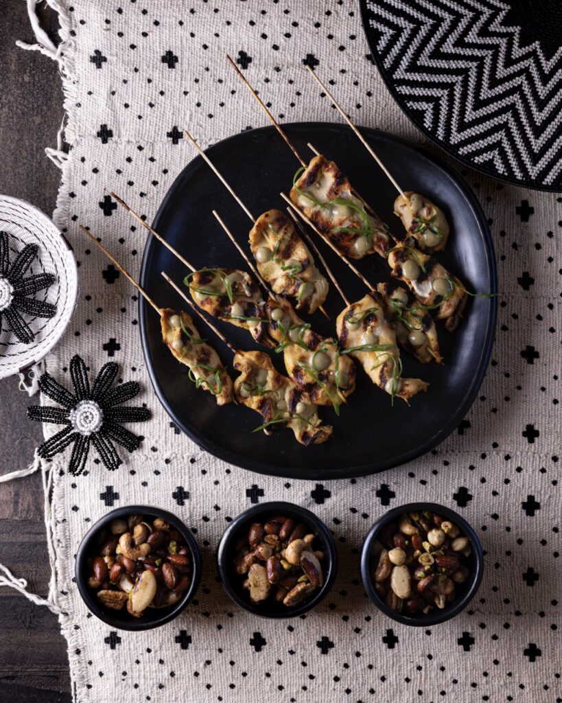 A large black dish has chicken skewers sitting on top and three small bowls below with nuts in them.