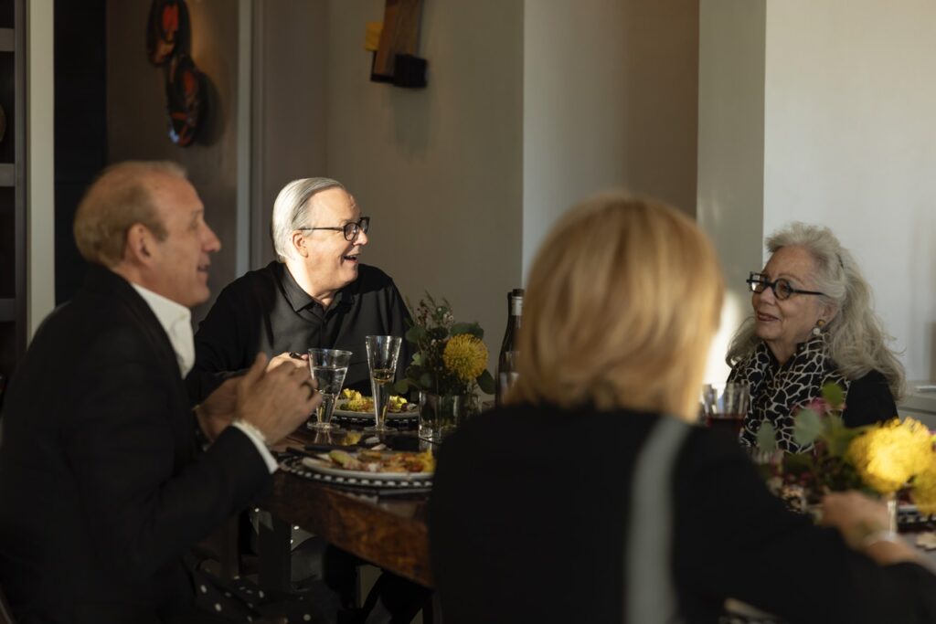 A group of people in black talk at a dinner table as the sun hits a man in his face who is stationed at the head of the table. 