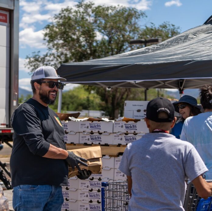 A man in a baseball hat and sunglasses holds a box with more boxes behind him as someone walks by with a shopping cart.