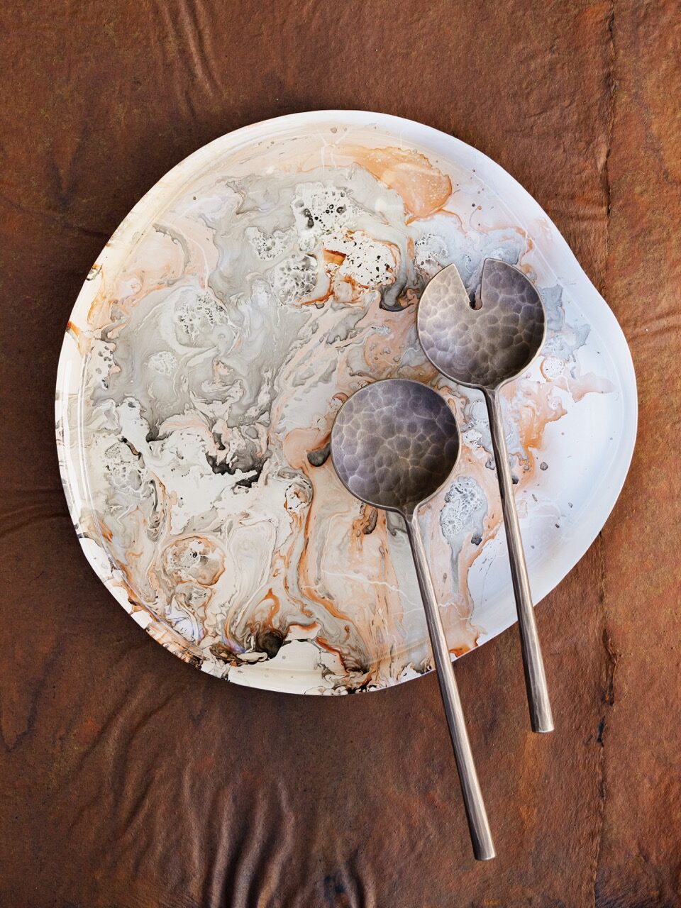 Two round circular spoons, one with a slice taken out of the head sit on an orange and grey marble plate, all on a brown table.