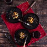 Three black bowls hold three servings of stuffing with forks inside each bowl and a red cloth below the bowls.