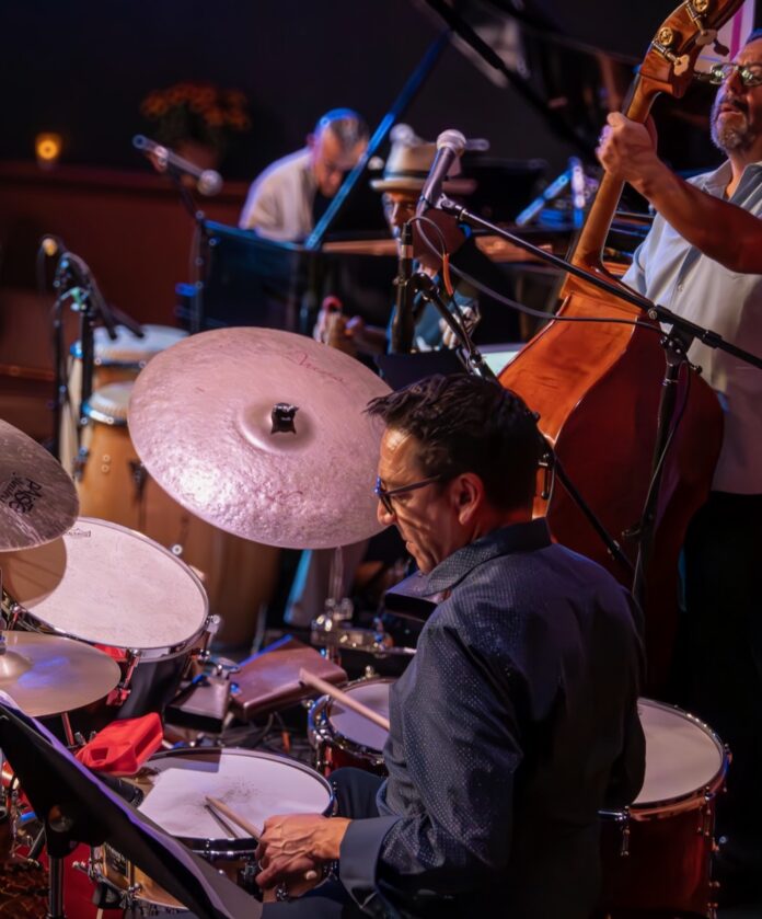 A man sits at a drumset while a man in the background plays upright bass at Unit B within Chocolate Maven.