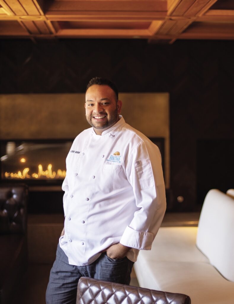 A chef stands in a white jacket with his hands in his pockets in front of a modern fireplace.