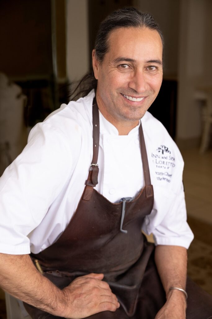 A chef sits against a table in a brown apron and white shirt.