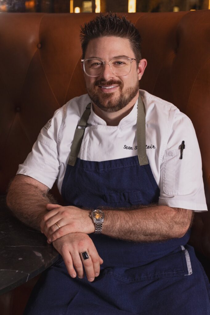 A chef stands with his arms crossed in front of him in a white shirt and blue apron.