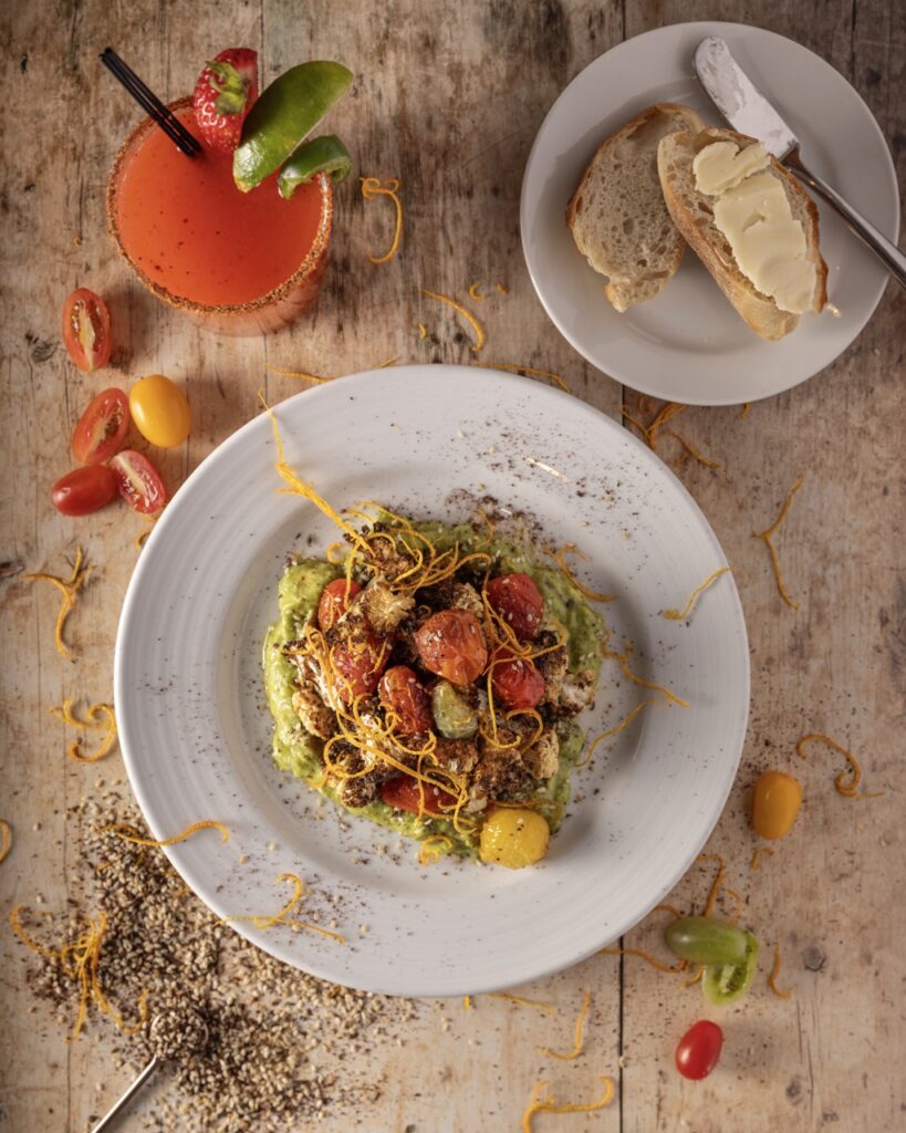 A white bowl full of a tomato salad with another small white plate with bread nearby and a red drink with a lime garnish.