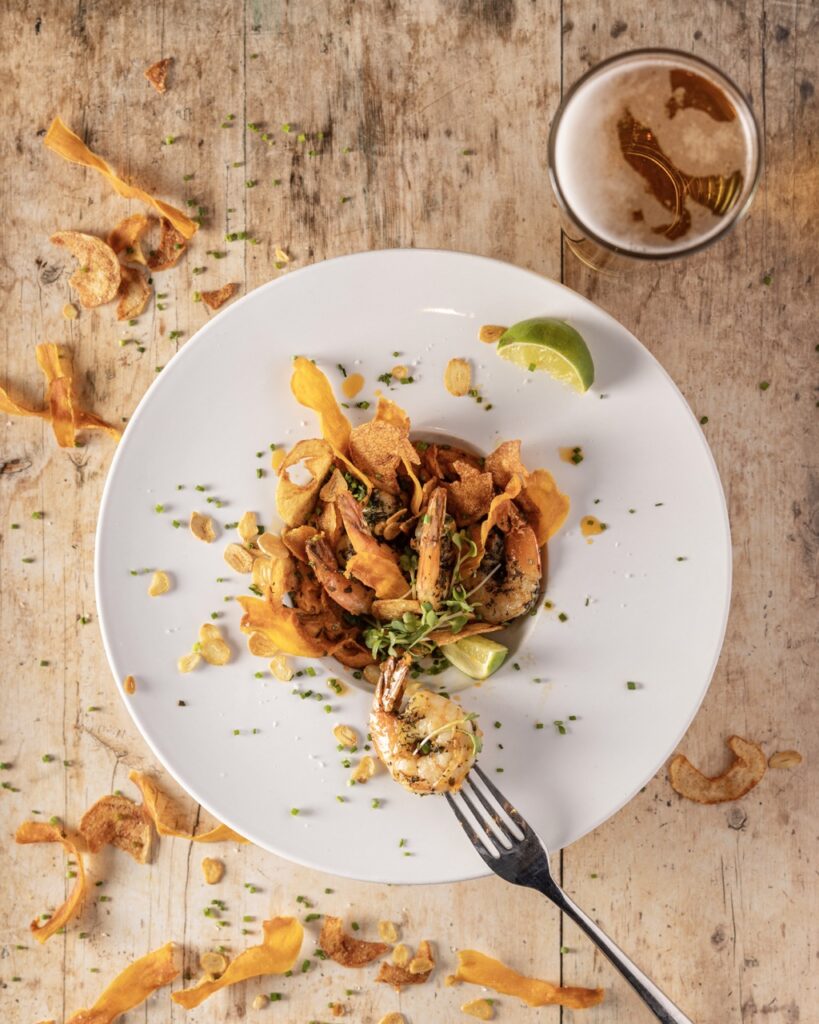 A shrimp pasta dish in a white bowl with a lime on the side and a beer at the top of the table.