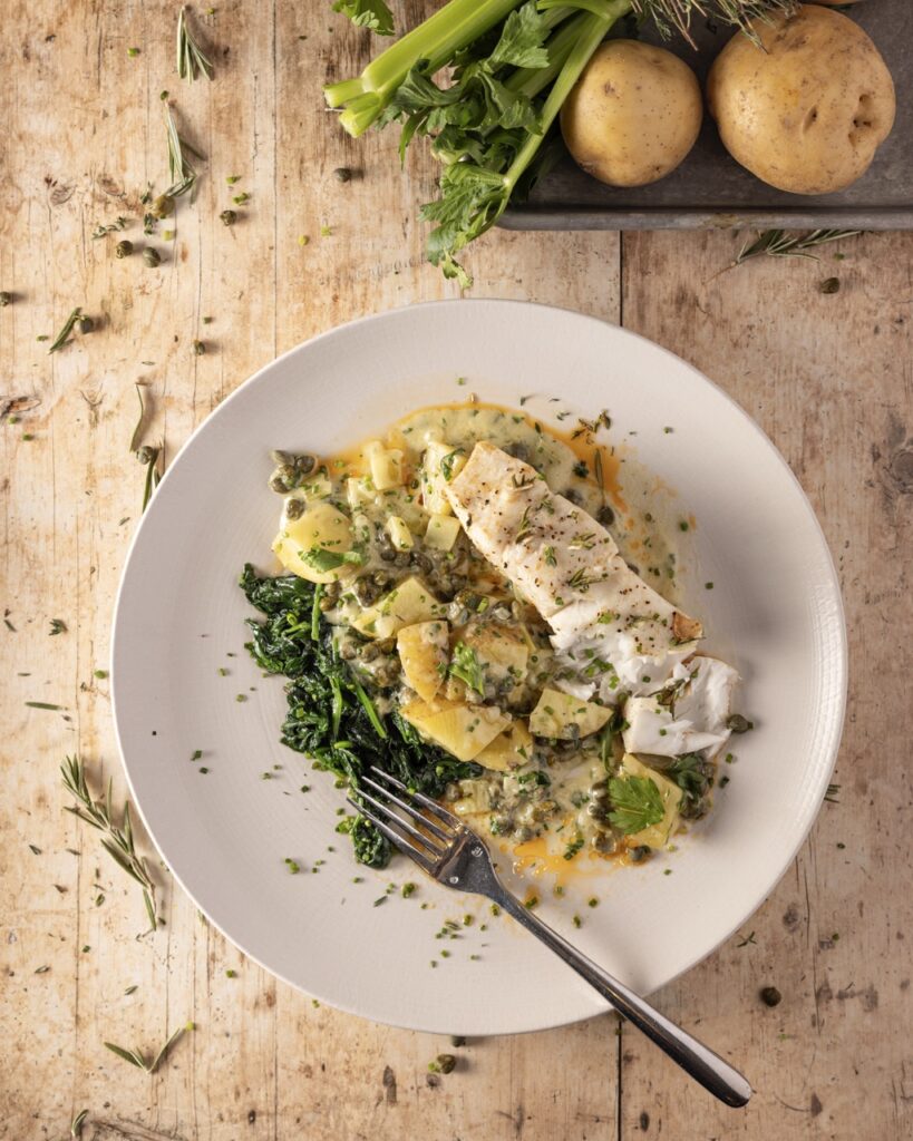 A chicken salad pasta sits on a white plate with herbs on the brown table below.