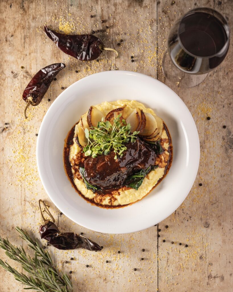 A white bowl of food holds a slice of meat on top of a bed of potatoes with chili peppers surrounding the plate.