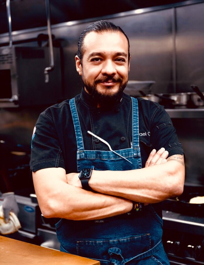 Chef Israel Castro stands in his kitchen with his arms crossed in front of his body and a blue apron on.