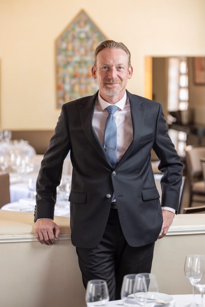 Rodolphe Louchart stands in the Compound dining room, leaning against a white table with a table set in front of him.